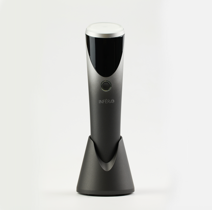 Sleek handheld INFERA device in a metallic finish, featuring a rounded treatment head, single control button, and charging stand, designed for targeted light therapy use.